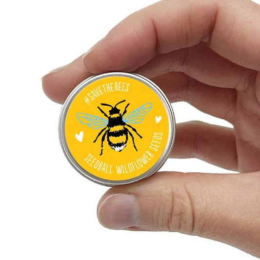 Hand holding a round yellow seedball tin with a bee design and text reading "#Savethebees Seedball wildflower seeds" on a white background