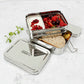 Stainless steel two tier lunch box with compartments containing fruits and bread and a separate tin with yoghurt on a light background.