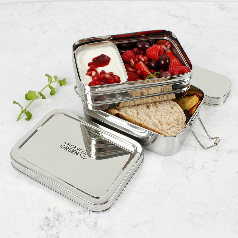 Stainless steel two tier lunch box with compartments containing fruits and bread and a separate tin with yoghurt on a light background.
