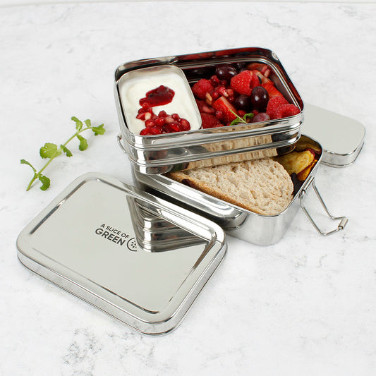 Stainless steel two tier lunch box with compartments containing fruits and bread and a separate tin with yoghurt on a light background.