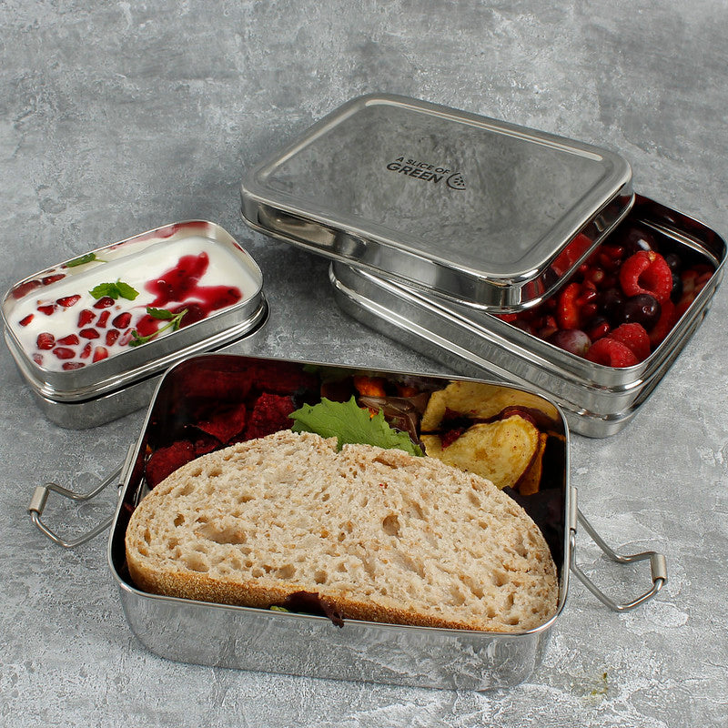 Stainless steel two tier lunch box with compartments containing fruits and bread and a separate tin with yoghurt on a grey background.