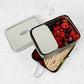 Stainless steel two tier lunch box with compartments containing fruits and bread and a separate tin on a light background.