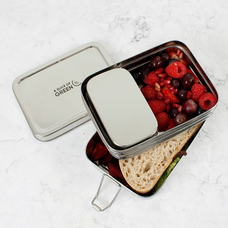 Stainless steel two tier lunch box with compartments containing fruits and bread and a separate tin on a light background.