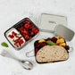 Stainless steel two tier lunch box with compartments containing fruits and bread and a separate tin with yoghurt on a light background, with a metal spork alongside
