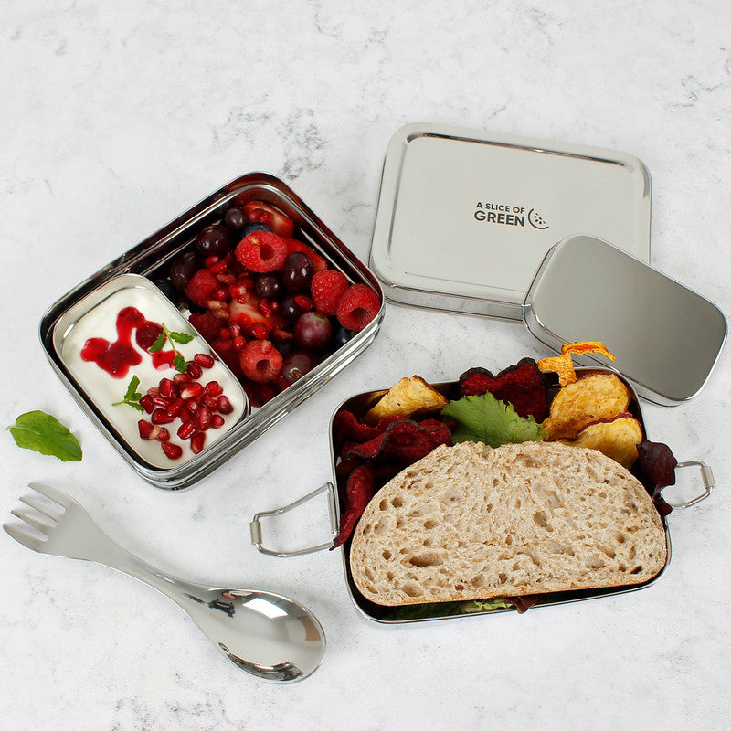 Stainless steel two tier lunch box with compartments containing fruits and bread and a separate tin with yoghurt on a light background, with a metal spork alongside