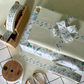 White paper tape with green floral design shown on the roll in a bamboo dispenser and applied to a wrapped gift