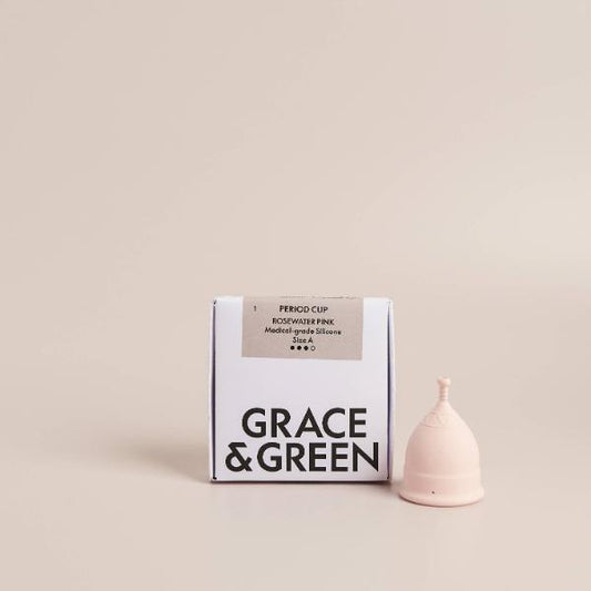 A reusable menstrual cup in pale pink sits beside its white and black Grace & Green packaging box on a warm beige studio background.
