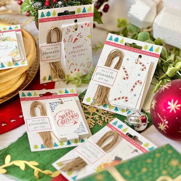 Four packages of craft twine gift tags arranged on a festive Christmas-themed surface with ornaments, greenery, and gift boxes. The packages are labeled 'Set of 10 planable gift tags' with holiday illustrations and tags in different designs.