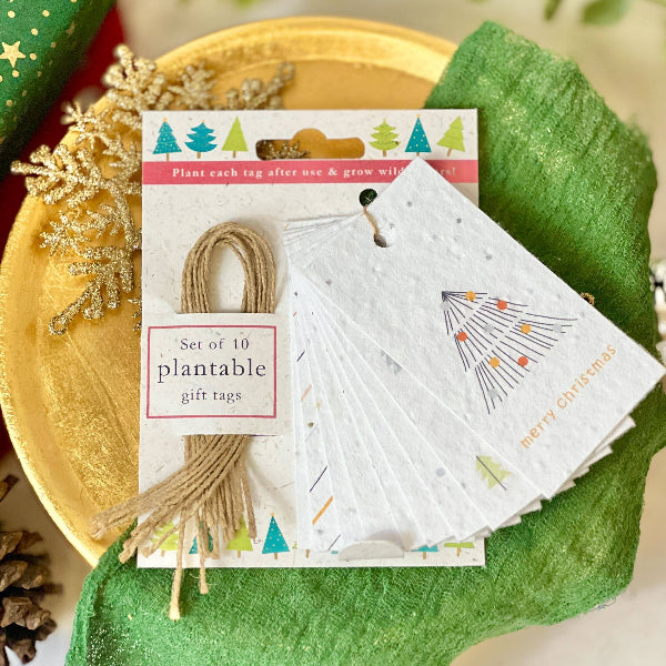 A set of 10 plantable gift tags displayed on a gold tray with green fabric. The tags are made from textured seeded paper, with one showing a simple black line drawing of a Christmas tree and gold 'Merry Christmas' text, and another showing a colourful tree pattern.