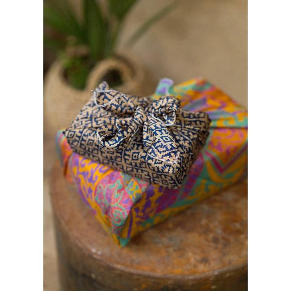 A close-up of a gift wrapped in sari fabric, placed on top of another brightly coloured wrapped package on a rustic wooden surface.