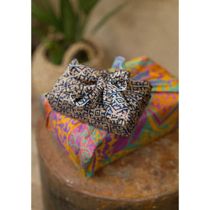 A close-up of a gift wrapped in sari fabric, placed on top of another brightly coloured wrapped package on a rustic wooden surface.