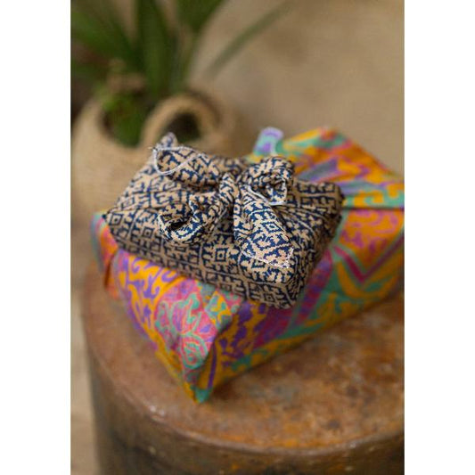 A close-up of a gift wrapped in sari fabric, placed on top of another brightly coloured wrapped package on a rustic wooden surface.