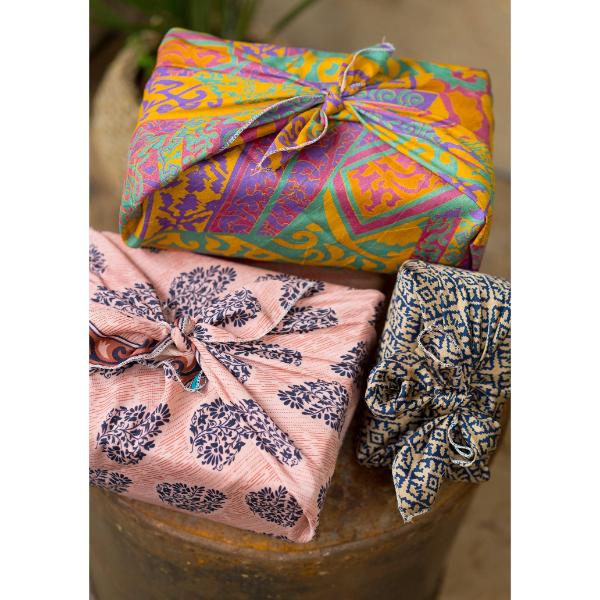 Three colourful packages wrapped in reusable recycled sari fabric, displayed on a rustic wooden surface. The top package has a bright yellow, orange, and purple geometric pattern.