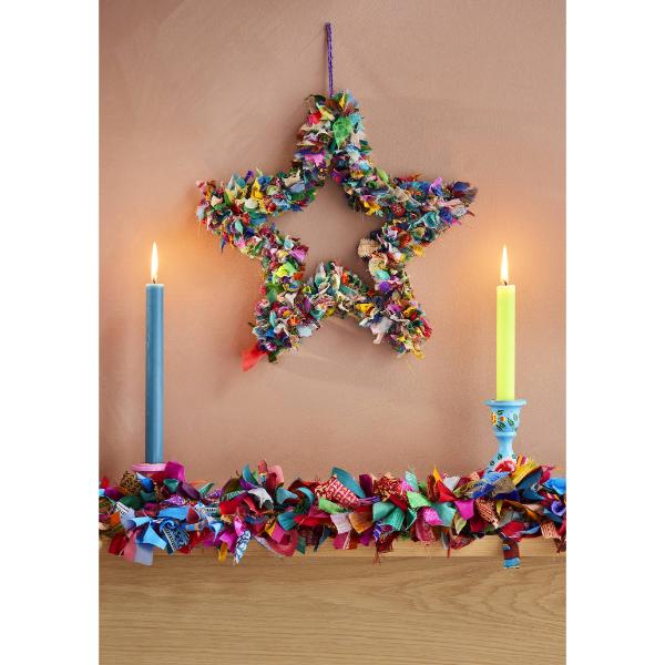 Recycled sari wreath using multicoloured sari remnants, shaped into a star design hanging on a wall next to two candles and some multicoloured sari tinsel along a mantelpiece