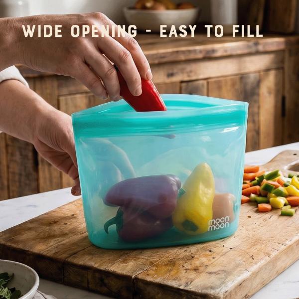 Person placing a red pepper into blue reusable food storage bag on a wooden cutting board with text reading "Wide opening - easy to fill"