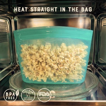 Popcorn in a teal silicone foot storage bag inside an open microwave with text reading "heat straight in the bag", and "BPA free, Platinum food grade silicone, FDA approved"