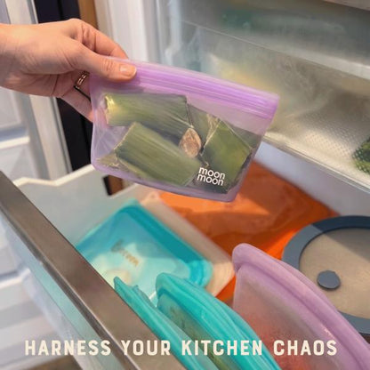 Hand holding a reusable food storage bag with 'Moonmoon' branding in front of an open freezer drawer with other bags inside and text reading "Harness your kitchen chaos"