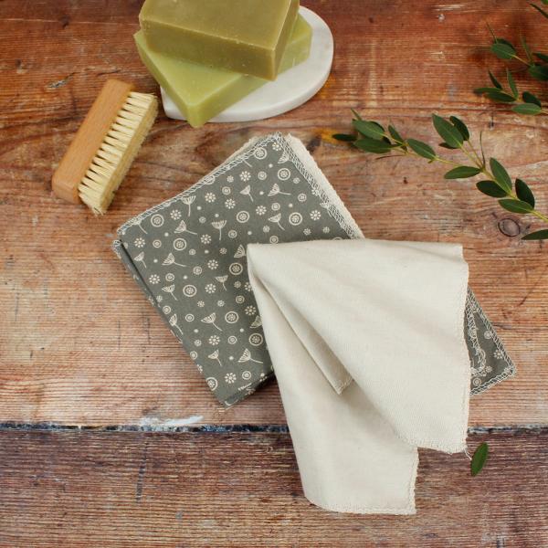 A set of reusable organic cotton wipes in a sage green and cream floral pattern, styled on a rustic wooden table with a wooden brush and green soap bars.