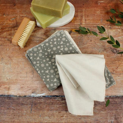 A set of reusable organic cotton wipes in a sage green and cream floral pattern, styled on a rustic wooden table with a wooden brush and green soap bars.