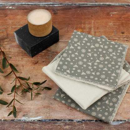 A stack of folded reusable wipes or cloths in muted grey and cream tones with a small repeating motif, arranged on a rustic wooden surface beside a candle and greenery.