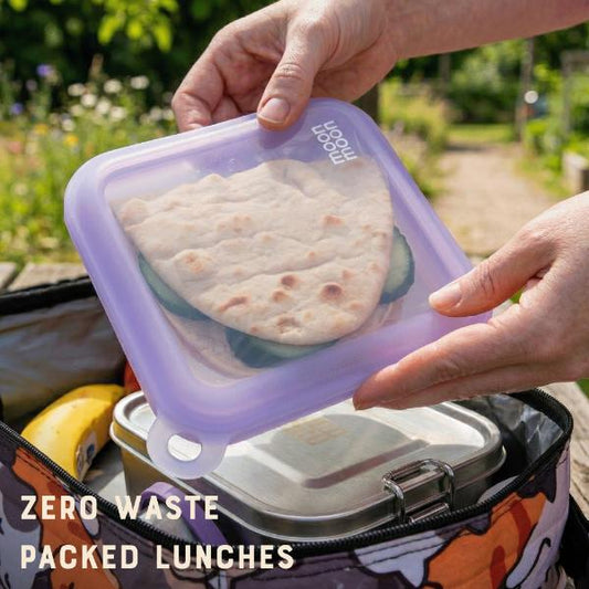 Person holding a purple silicone reusable sandwich case with flatbread sandwich, set against a garden background.