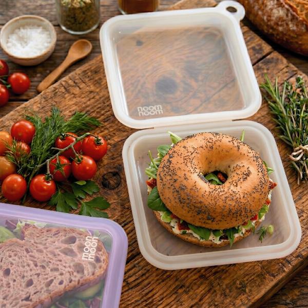 Bagel sandwich in a reusable sandwich case on a wooden table with fresh ingredients.