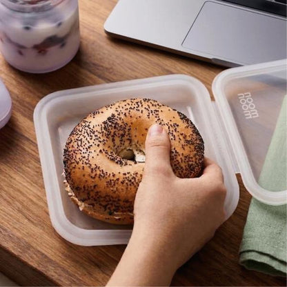 Person holding a bagel in a silicone sandwich case on a wooden table with a laptop and yogurt.