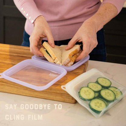 Person preparing a sandwich with a container of cucumber slices on a wooden cutting board putting sandwiches into reusable silicone sandwich case and text reading "Say goodbye to cling film"