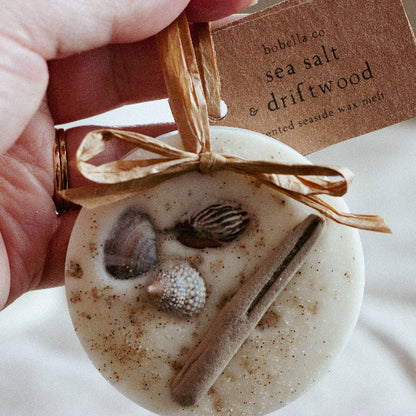 A hand holding a acented home decoration in 'beach and coastal' scent, wax melts with shells and driftwood embedded in the wax, with a raffia hanging loop