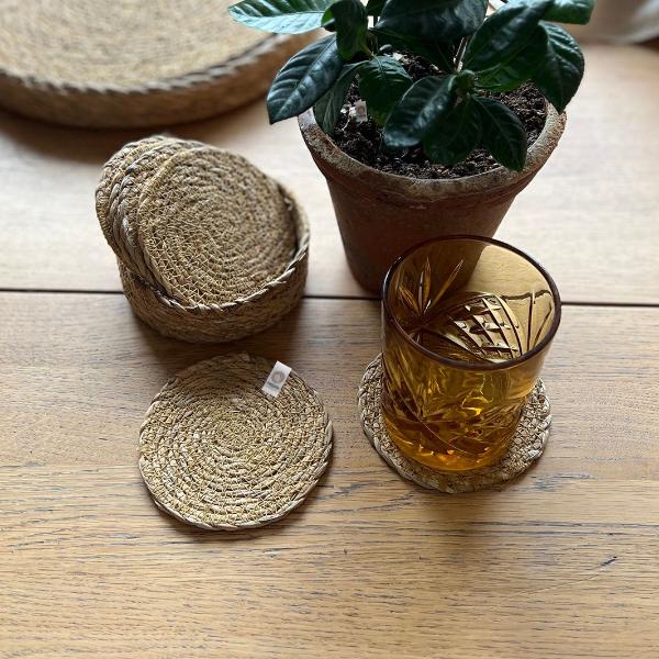 Set of 6 natural seagrass coasters with a holder, with a glass on one, alongside a potted plant