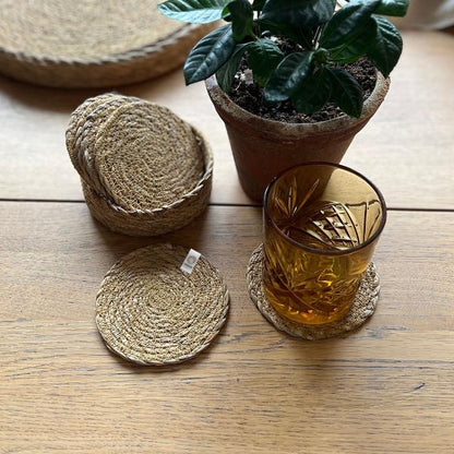 Set of 6 natural seagrass coasters with a holder, with a glass on one, alongside a potted plant