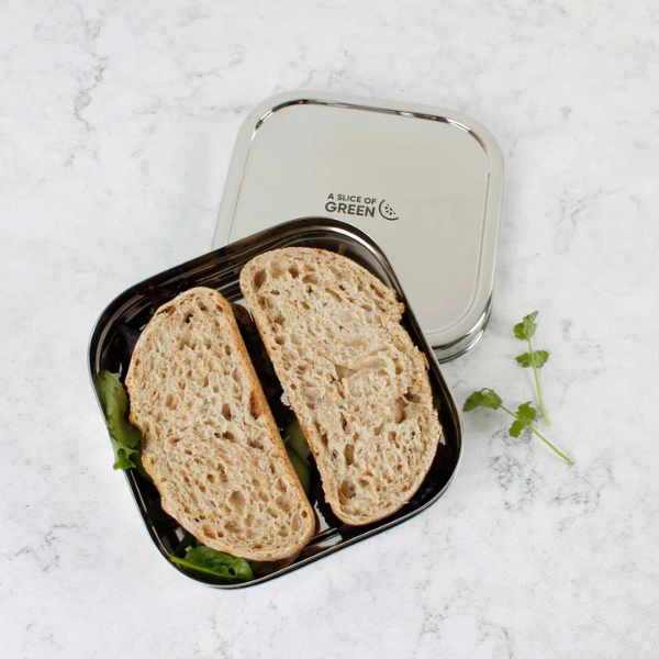 Stainless steel sandwich lunchbox shown open with sandwich inside, 'A Slice of Green' on lid and sitting on a marble surface.