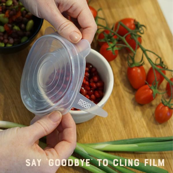 Person using a silicone food cover on a container with tomatoes and green onions on a wooden surface and text reading "say goodbye to cling film"