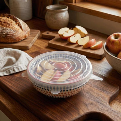 A bowl with a silicone lid and containing sliced apples sits on a wooden cutting board in a kitchen setting, with sliced apples, bread, and ceramic vessels in the background.