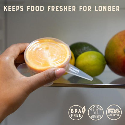 Hand holding half an orange covered with a silicone lid, surrounded by fruits, with text 'Keeps food fresher for longer' and certification logos.