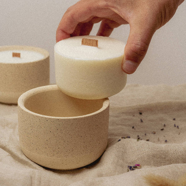Hand lifting a soy wax candle into a reusable candle pot on a textured surface.