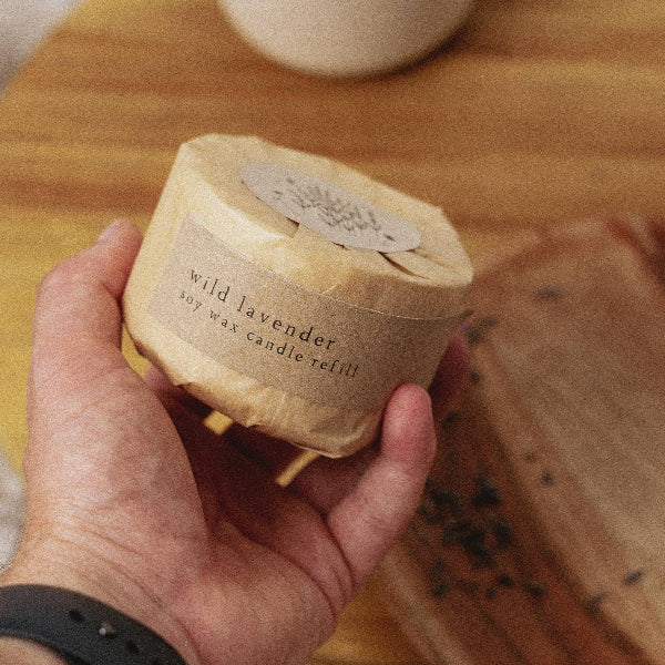 Hand holding a wax candle refill labeled 'wild lavender' on a wooden surface.