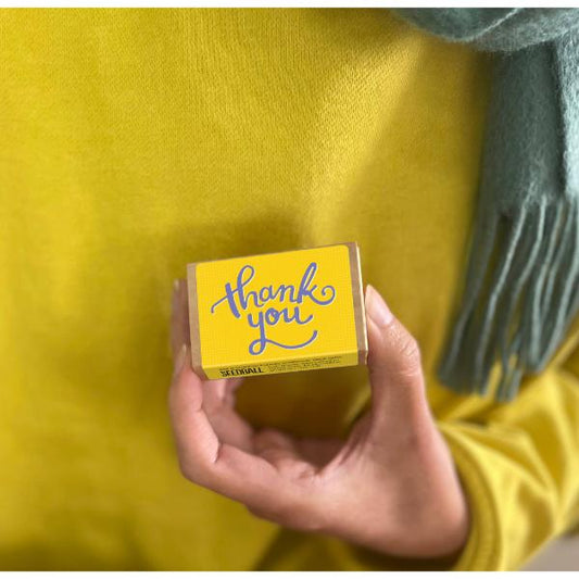 Hand holding a yellow seedball box block with 'thank you' text against a yellow jumper background