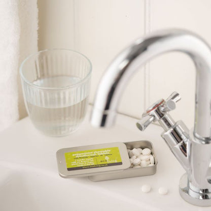 Toothpaste tablets in a tin, with fluoride on a bathroom sink next to tap and glass of water, shown open with tablets inside