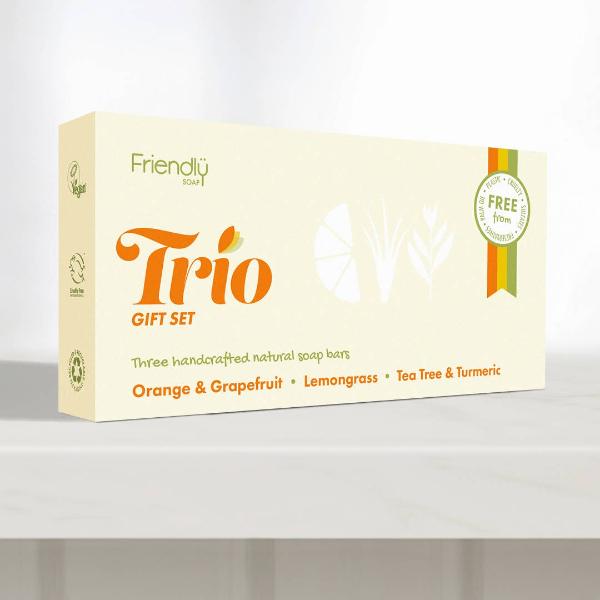 A cream-colored gift set box for 'Friendly Trio' natural soaps, featuring orange and green text and illustrations, displayed on a white shelf against a light grey background.