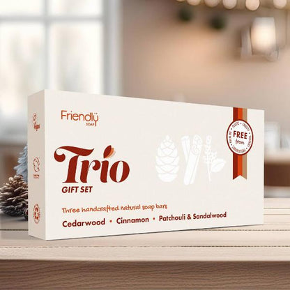 A white box of Friendly Soap Trio Gift Set, featuring natural soaps: Cedarwood, Cinnamon, and Patchouli & Sandalwood. The box is displayed on a wooden surface with a blurred warm indoor background.