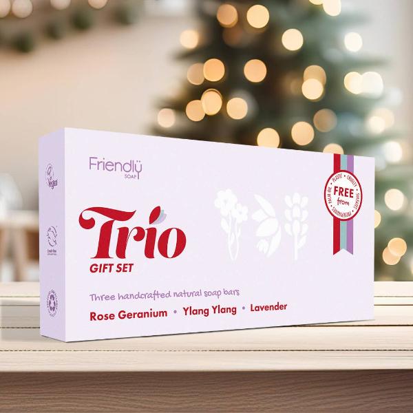A white rectangular gift set box labeled 'Friendly Soap Trio GIFT SET' with red and purple text, placed on a wooden surface with blurred warm lights in the background.