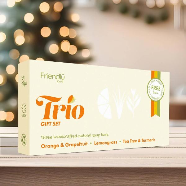 A cream-colored soap gift set box labeled 'Friendly Soap Trio Gift Set' with orange and green text, placed on a wooden surface with a blurred background.