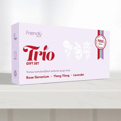 A light lavender box for the Friendly Trio Gift Set, containing three handcrafted natural soap bars (Rose Geranium, Ylang Ylang, and Lavender). The box features red and purple text and illustrations, set against a clean white background.