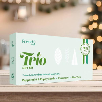 A boxed soap gift set labelled 'Friendly soap Trio' sits on a wooden surface with a softly blurred background. The box is pale mint green with dark green text and small white leaf illustrations.