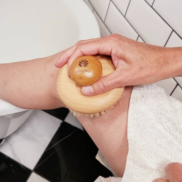 Person using a wooden body brush on their leg with a ecojiko logo in a bathroom setting.