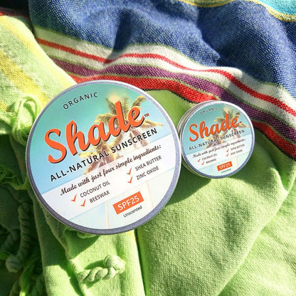 100ml and 15ml tins of Shade all-natural sunscreen with a colourful label on a white background, shown on blue and green fabric