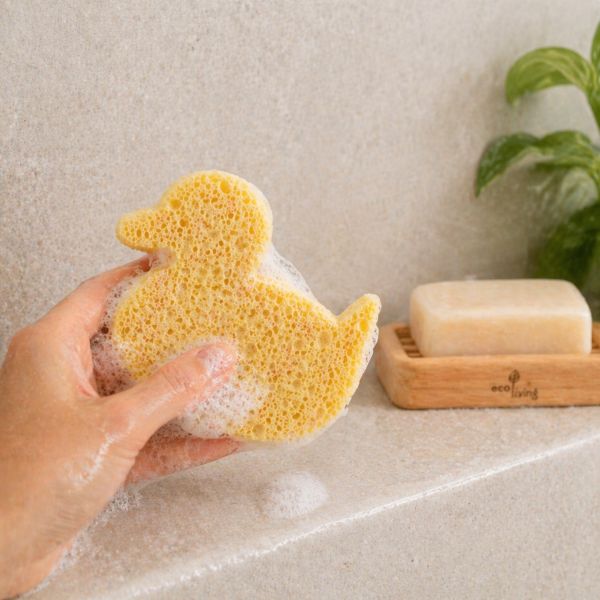 Hand holding yellow duck-shaped home compostable sponge with soap suds, next to a bar of soap on a wooden dish.
