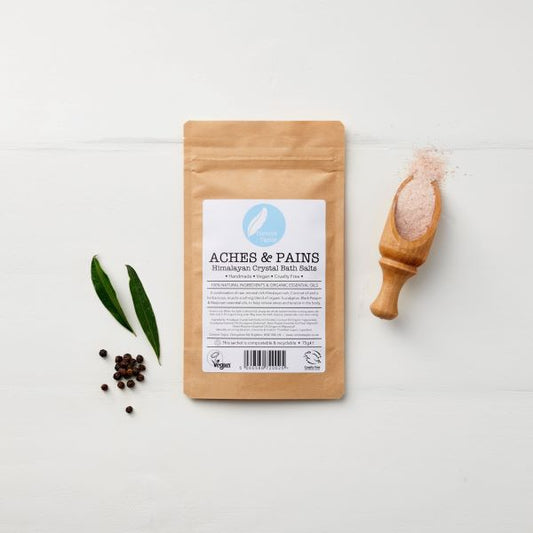 A kraft paper pouch of Corinne Taylor Aches and Pains Himalayan Bath Salts, on a white background with a wooden scoop of pink bath salt crystals, alongside bay leaves and peppercorns.